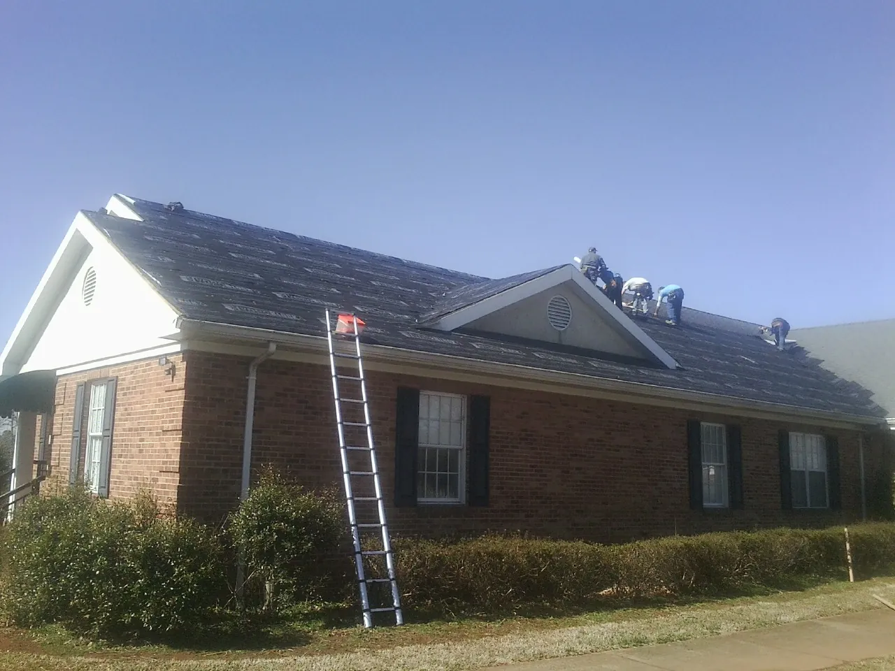 Crew tearing shingles off a brick ranch roof with a ladder set — Metal Roof Repair work by our crew in Eagle Bend