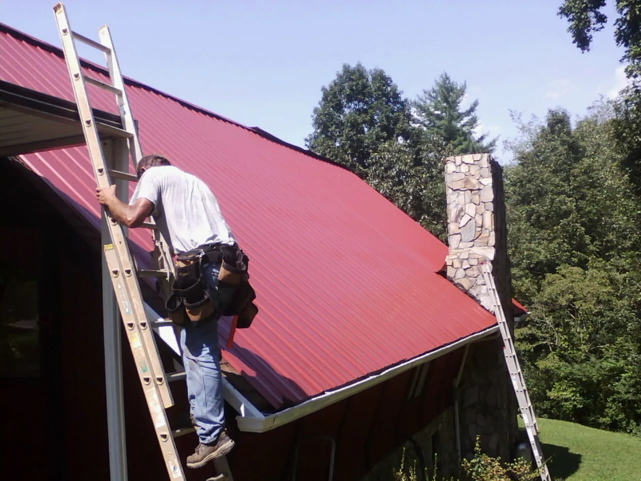 Roofer on a ladder beside a red metal roof and stone chimney — Soffit & Fascia Repair by Roofing Brothers in Eagle Bend